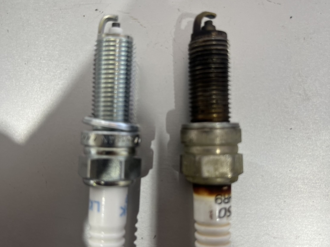交換前後のスパークプラグを並べた比較写真。右は汚れやカーボンが蓄積された使用済みプラグ、左は光沢のある新品プラグ