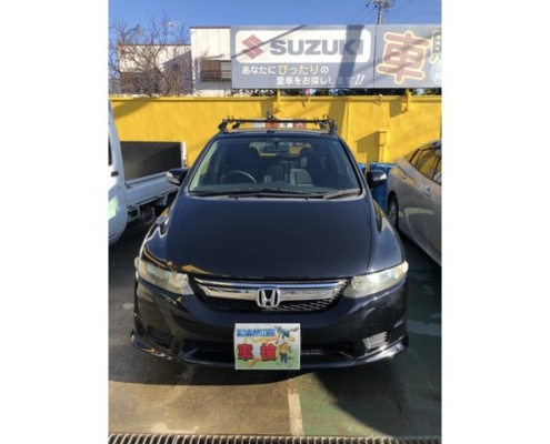 愛知県江南市の整備工場にて車検整備を行ったホンダ オデッセイ RB型の正面写真。屋外駐車場にて撮影され、背景にはスズキの看板と黄色い外壁が見える。ナンバープレートには“車検”の文字と整備士のイラストが描かれている。