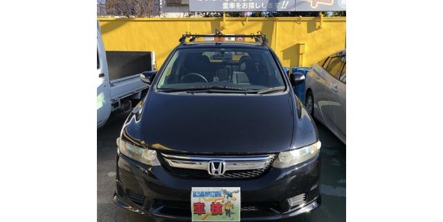 愛知県江南市の整備工場にて車検整備を行ったホンダ オデッセイ RB型の正面写真。屋外駐車場にて撮影され、背景にはスズキの看板と黄色い外壁が見える。ナンバープレートには“車検”の文字と整備士のイラストが描かれている。